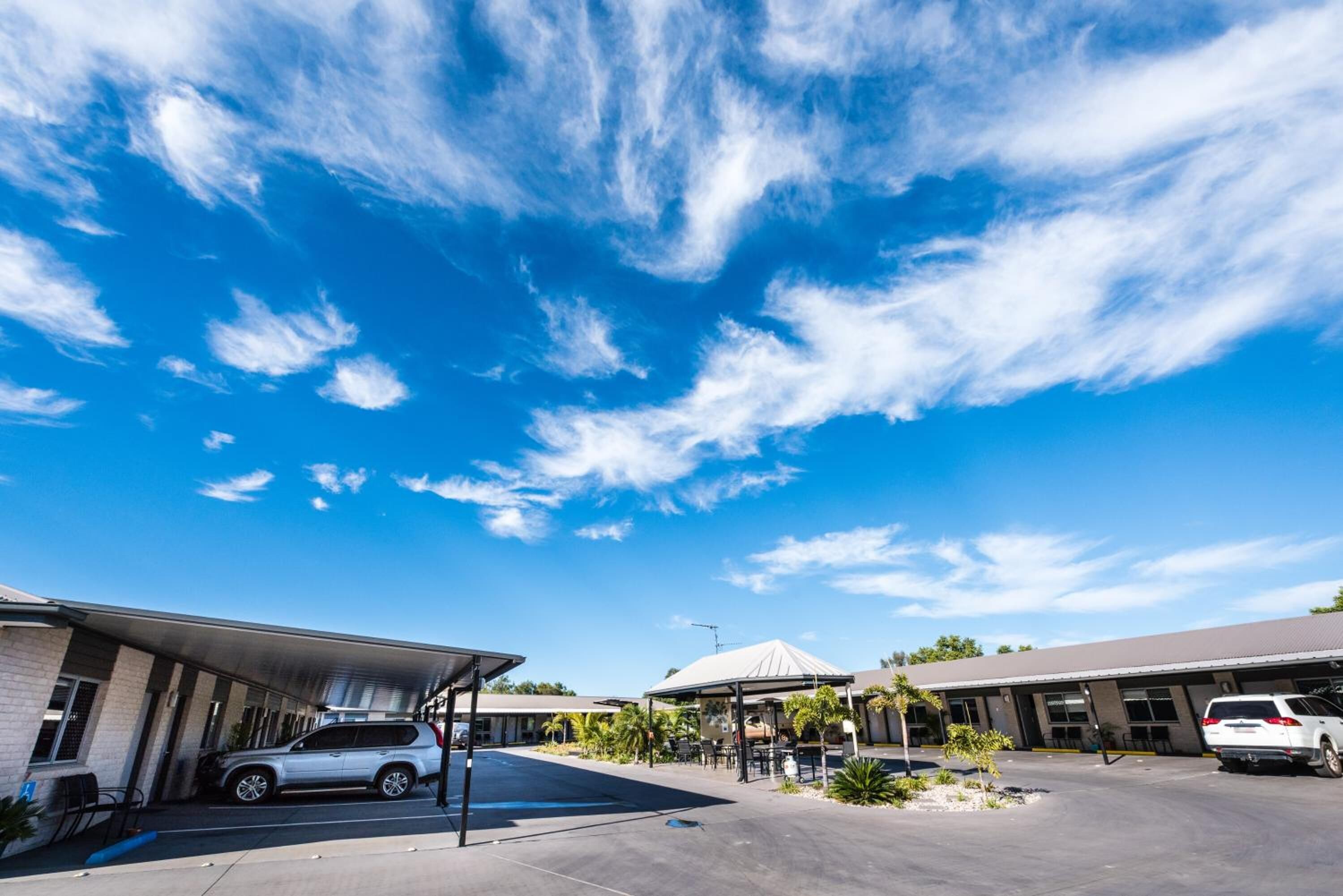 Un estacionamiento con área de lavado de coches techada, un edificio de techo plano y un cielo azul despejado con algunas nubes.
