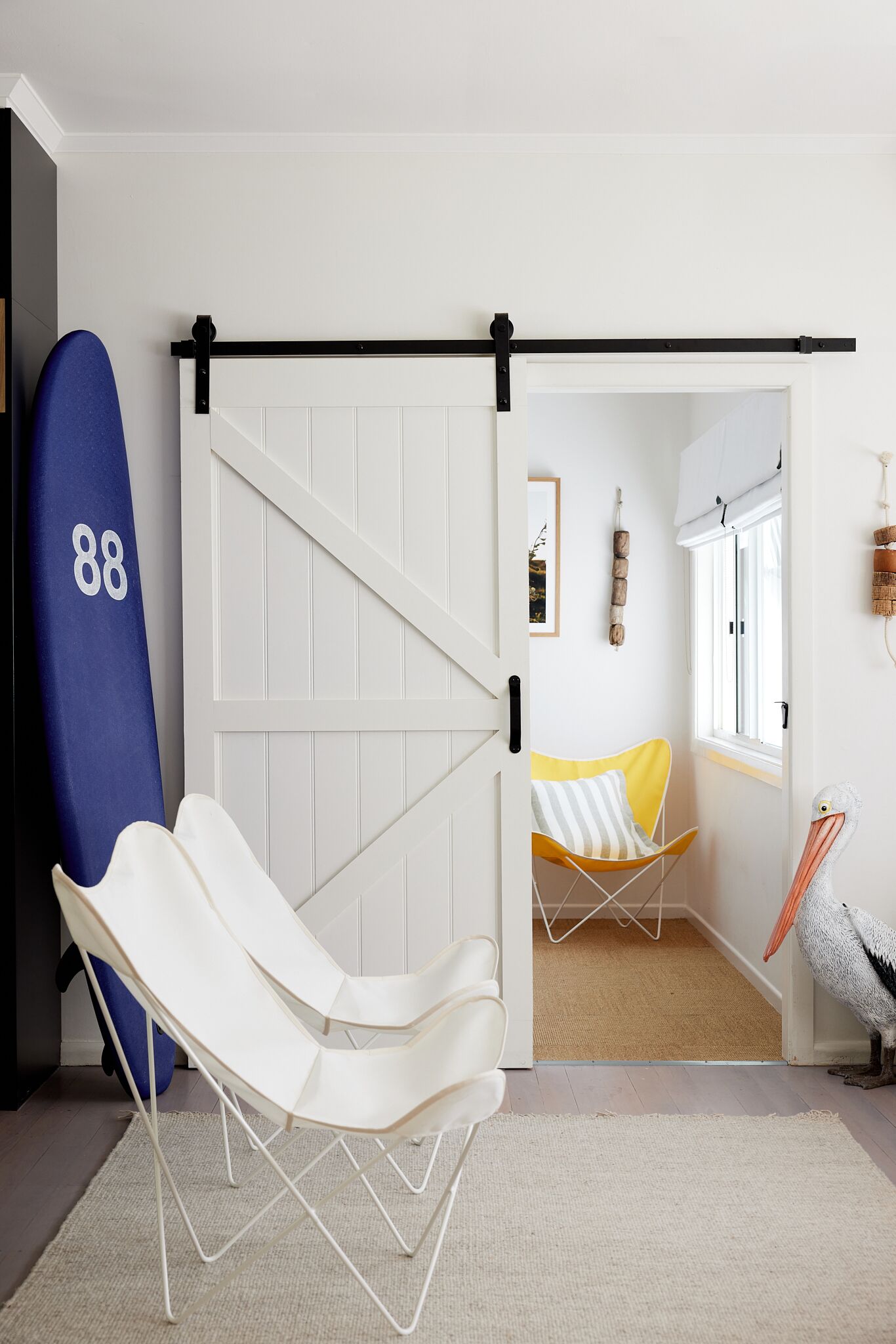 A room with a white chair, a blue surfboard, a yellow chair, and a pelican sculpture.