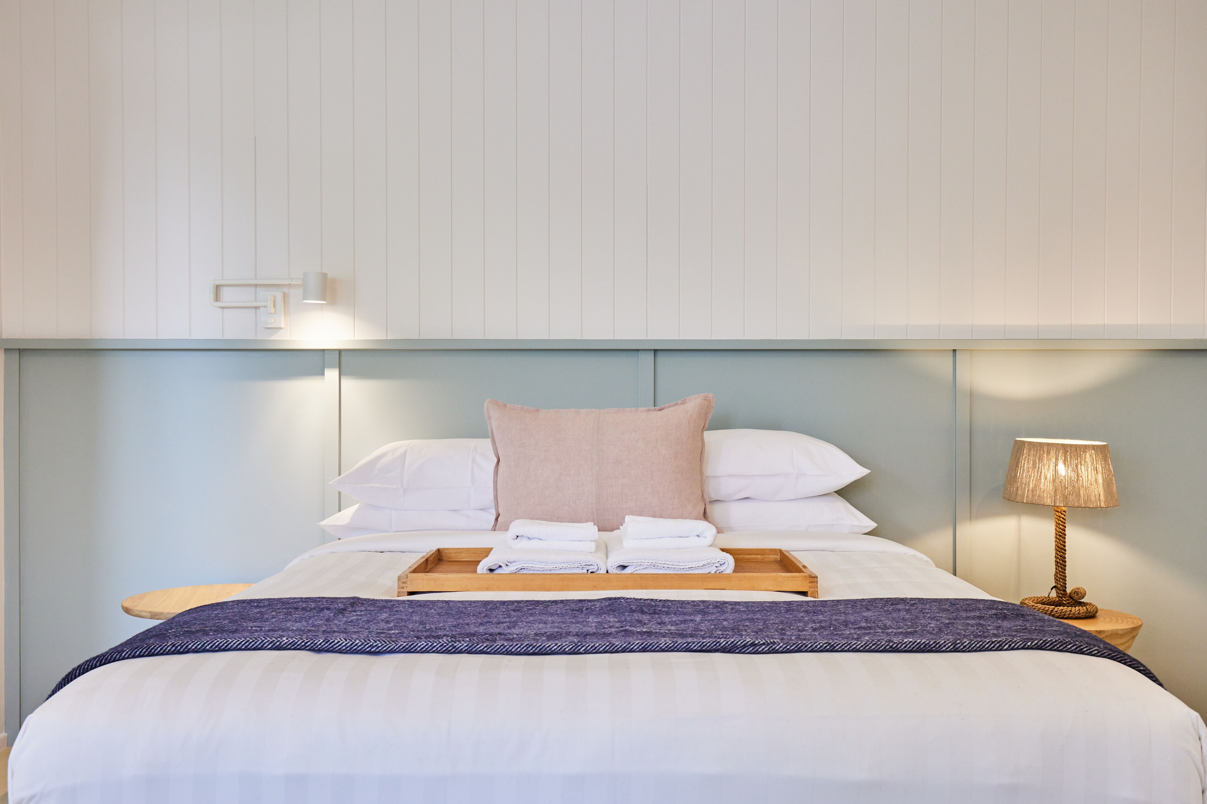 Un lit bien fait, avec une tête de lit bleue, une lampe de chevet et un plateau sur lequel sont pliées des serviettes.