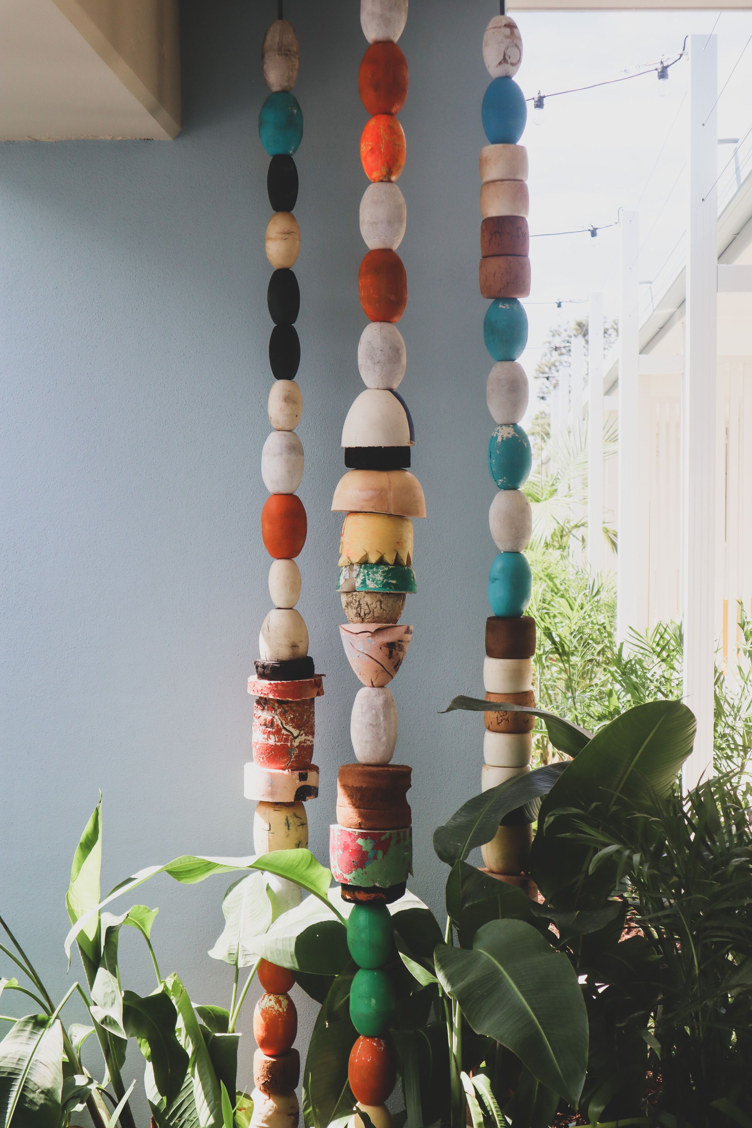 Una composición vertical de objetos de diversos colores y formas, posiblemente elementos decorativos, frente a una pared de tonos claros con una ventana y follaje en primer plano.