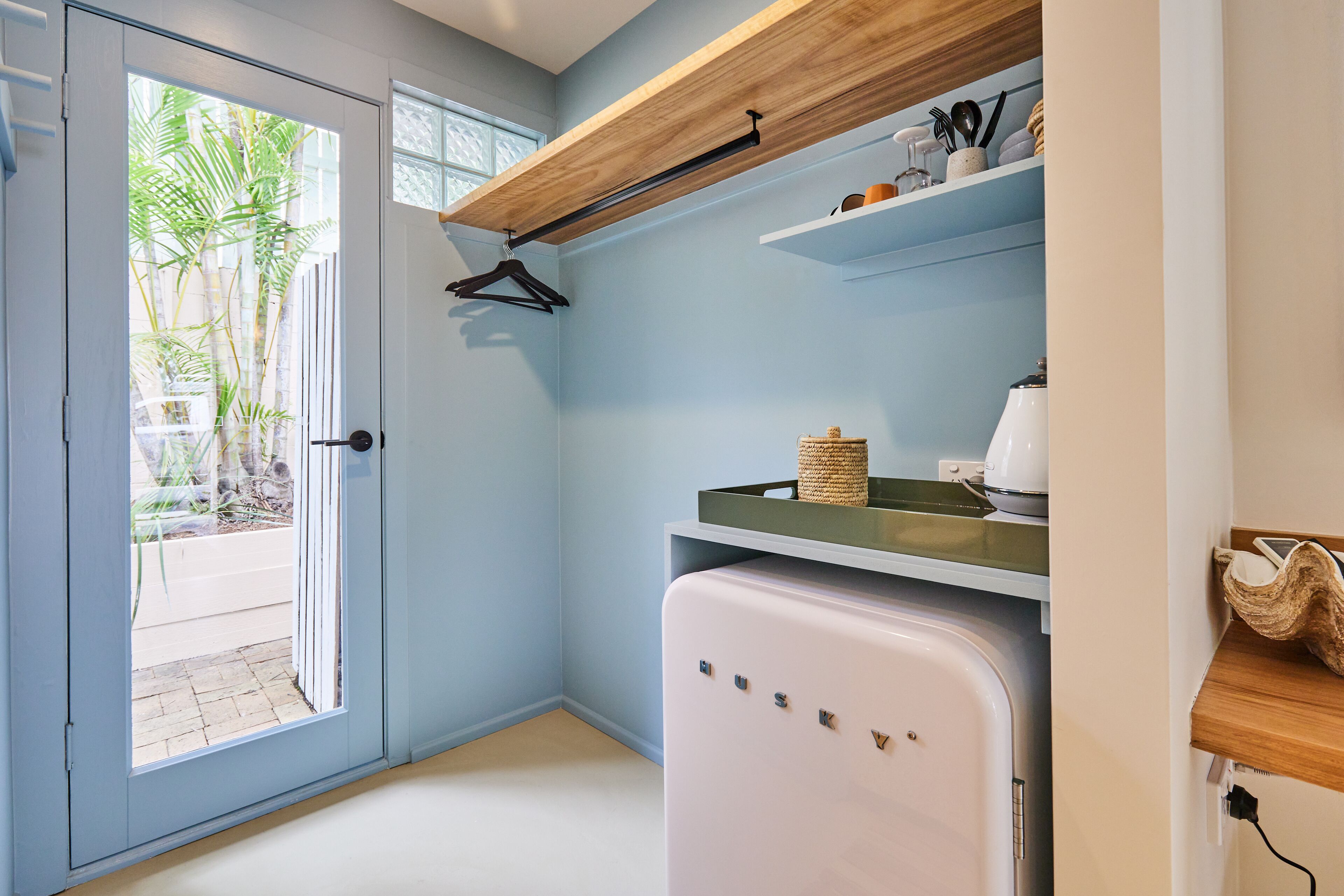 A compact kitchen with a white refrigerator, wooden shelves, and a hanging coat rack.