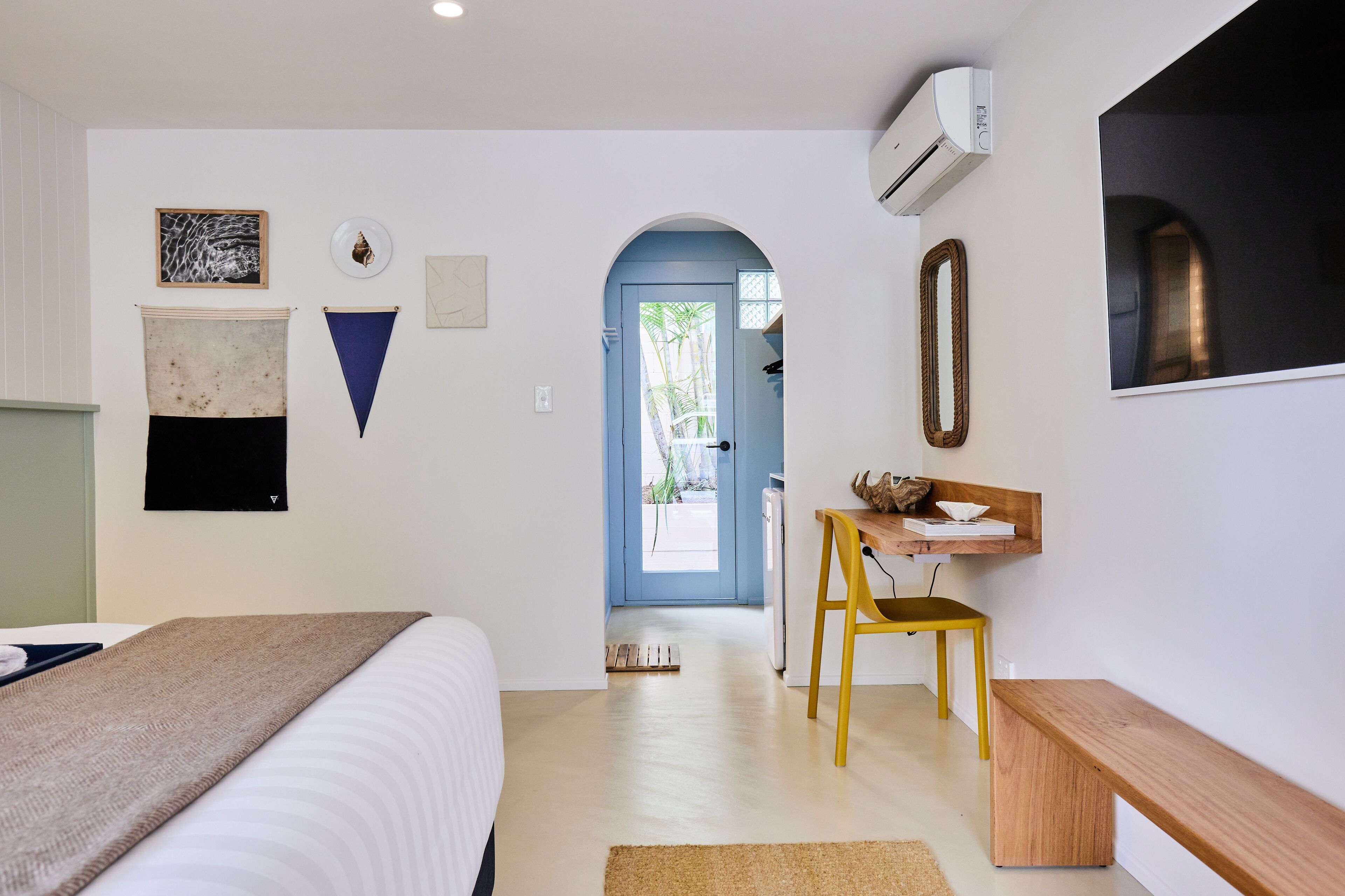 Una habitación de hotel moderna con cama, escritorio, silla y banco. Se observa una puerta que da a un balcón con vistas al exterior.