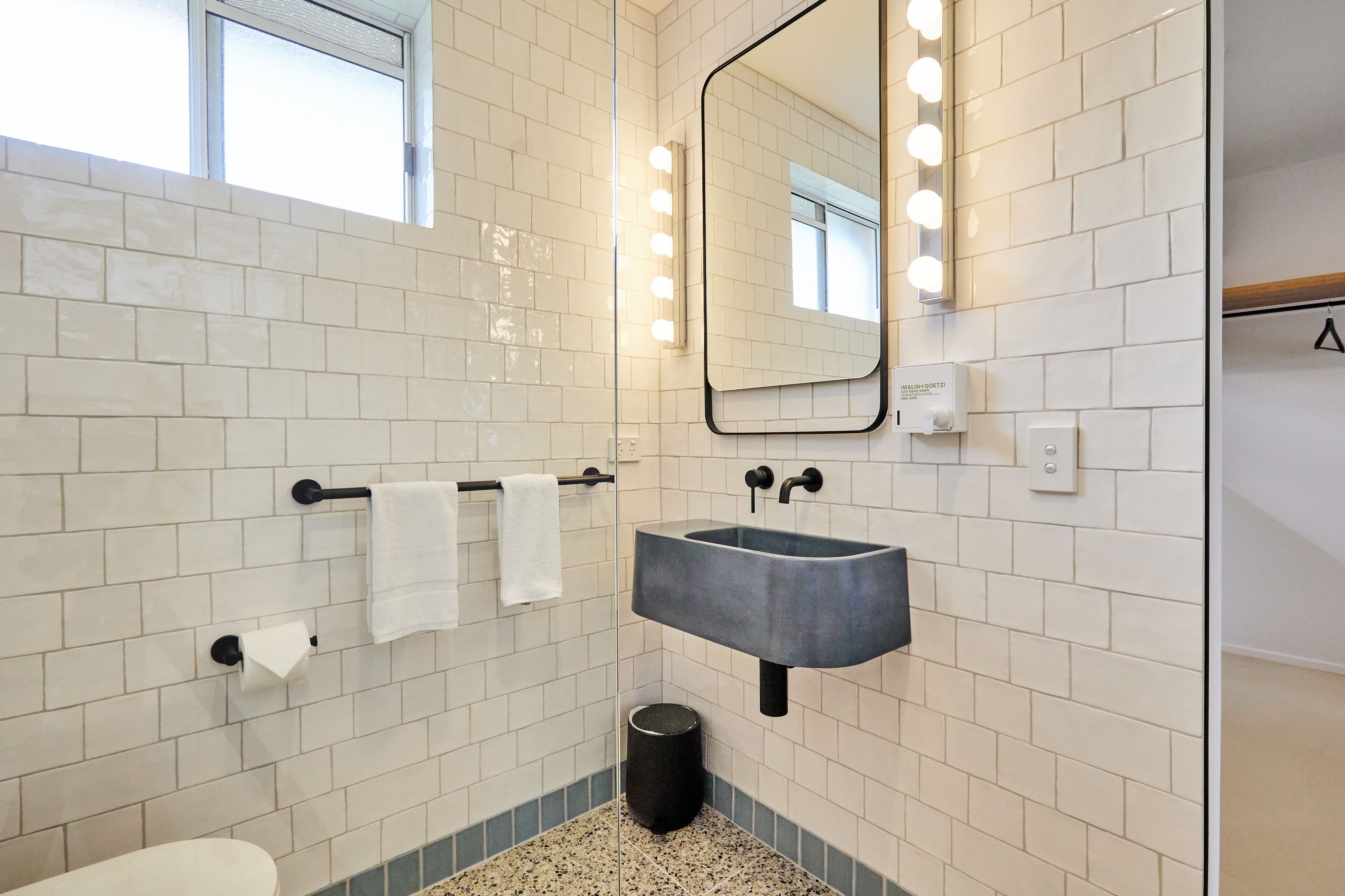 A bathroom with white subway tiles, a black sink, a mirror with lights, and a towel rack.