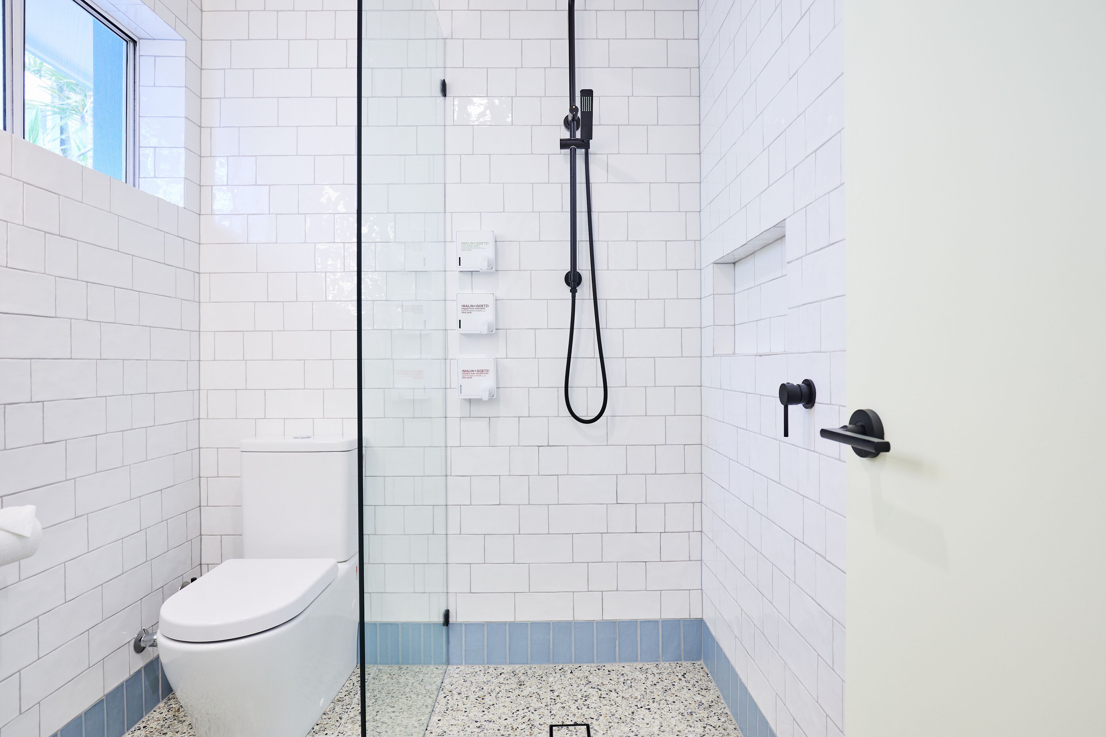 A bathroom with white tiled walls, a white toilet, a shower with a black hose, and a blue-tiled floor.
