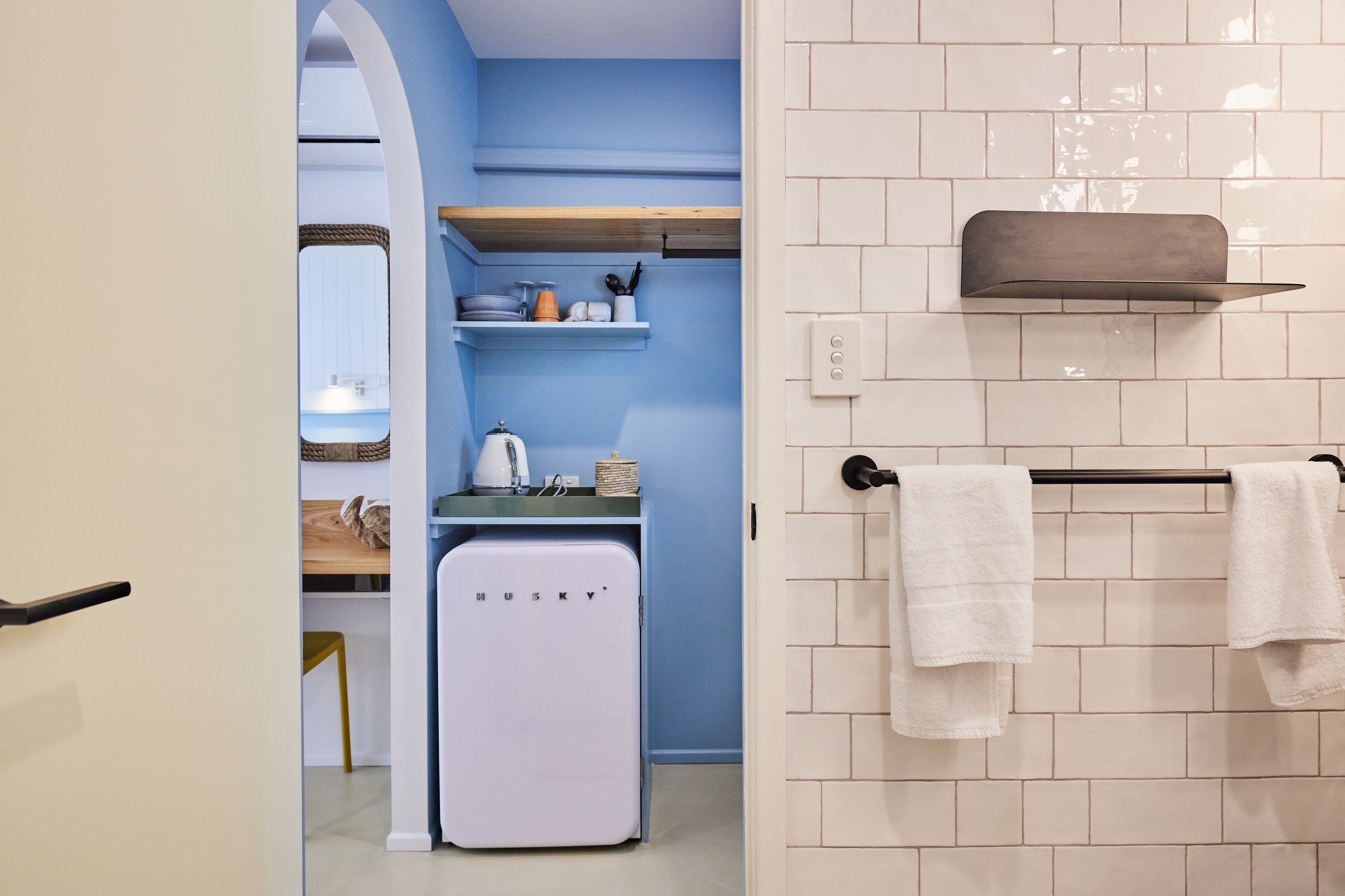 A modern kitchen with a compact refrigerator, a sink, and a wooden shelf above it.