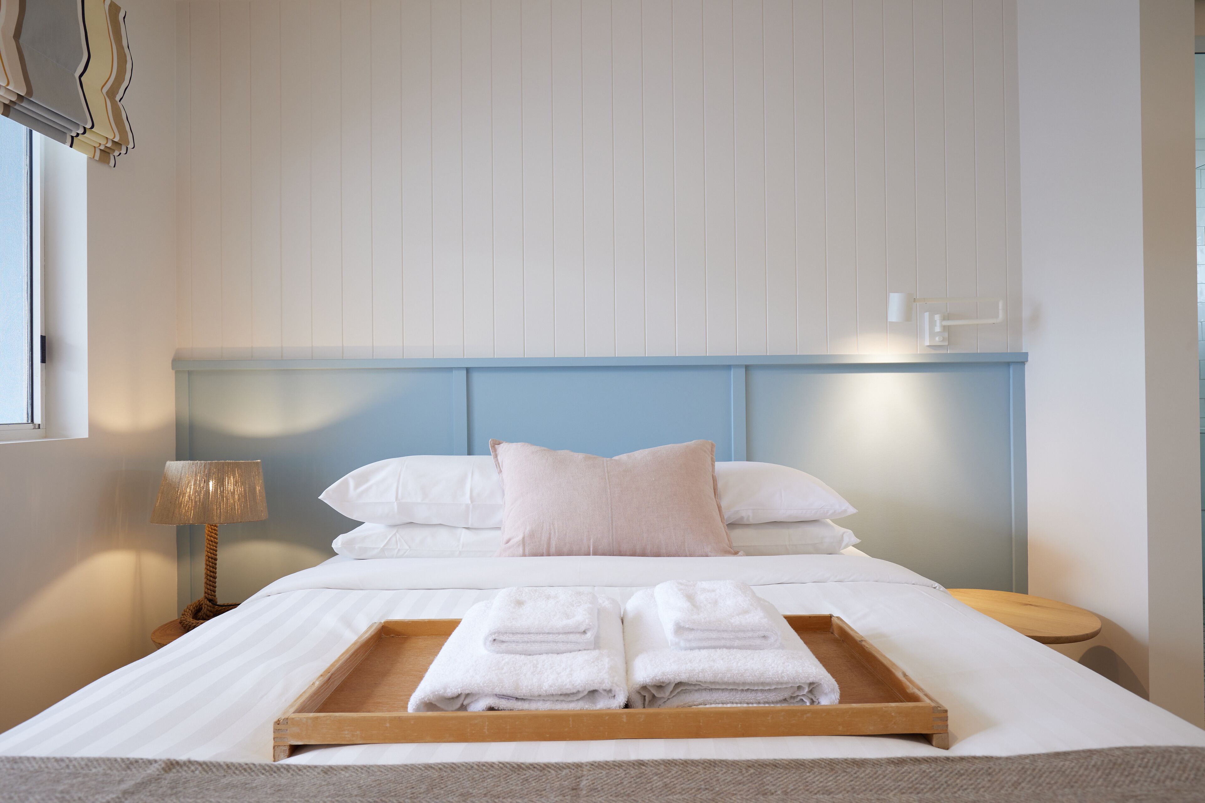 A neatly made bed with a wooden tray of folded towels, a bedside lamp, and a wall-mounted light fixture.