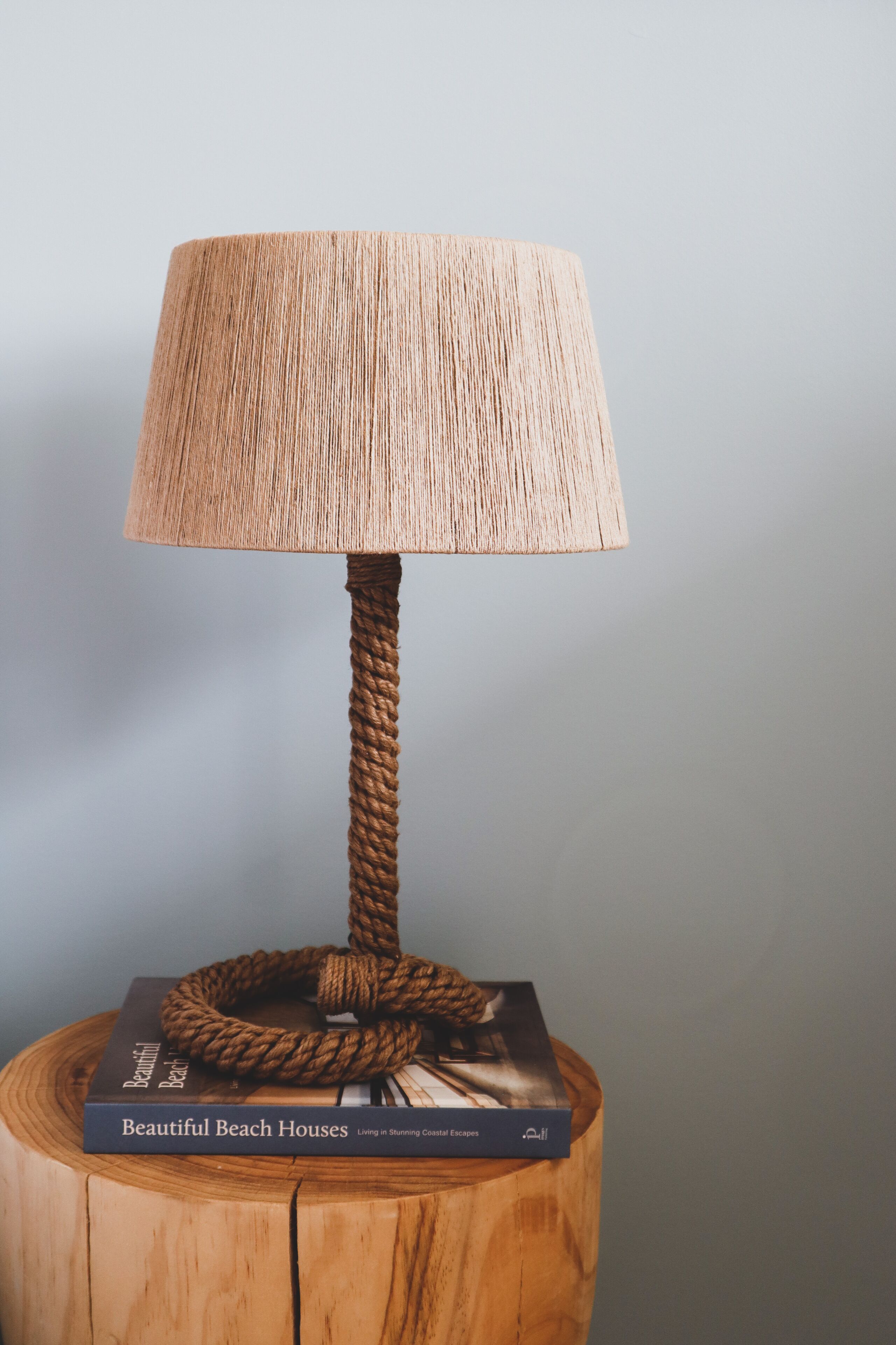 Une lampe dotée d’un pied en corde et d’un abat-jour tissé, posée sur un tronc d’arbre, sur lequel se trouve un livre intitulé « Belles maisons de plage ».