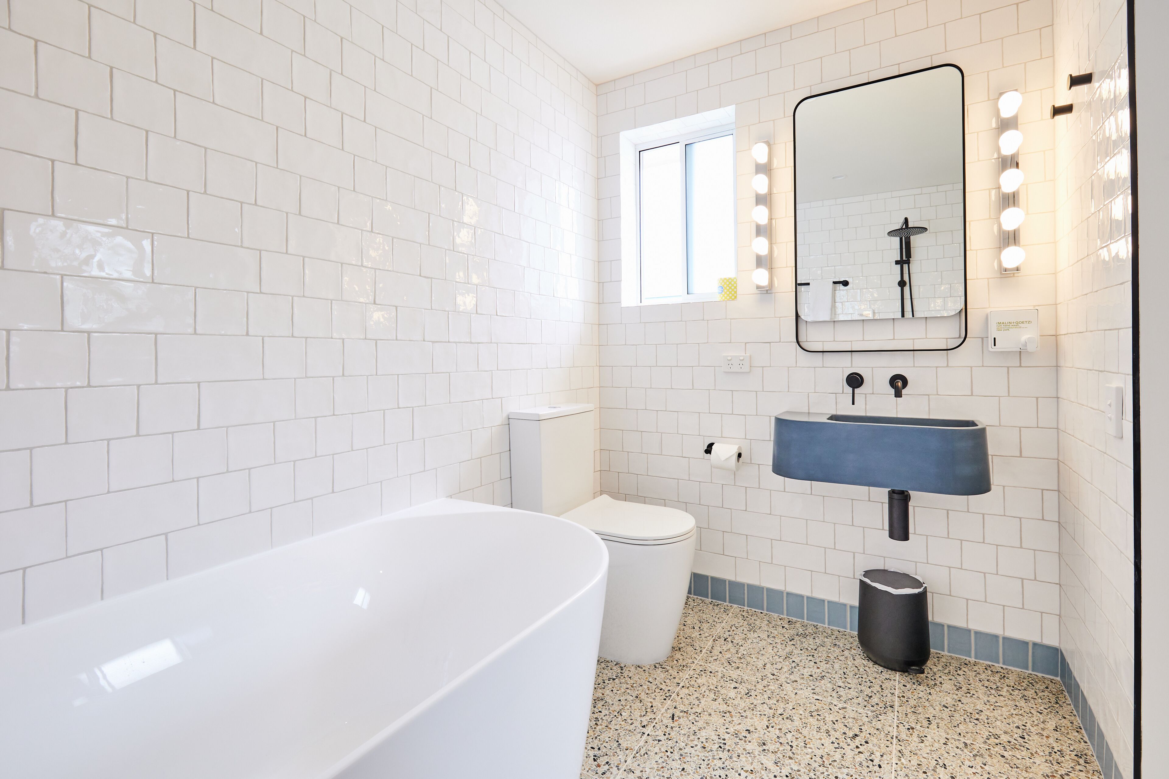 Een badkamer met witte metrotegels, een witte badkuip, een blauwe wastafel, een toilet en een spiegel met verlichting.