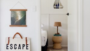 A hallway with a white door leading to a bedroom. There is a wall-mounted shelf with a framed picture, a wall-mounted lamp, a basket, and a bag with the word "ESCAPE" on it. - Blue Water Motel (Kingscliff)