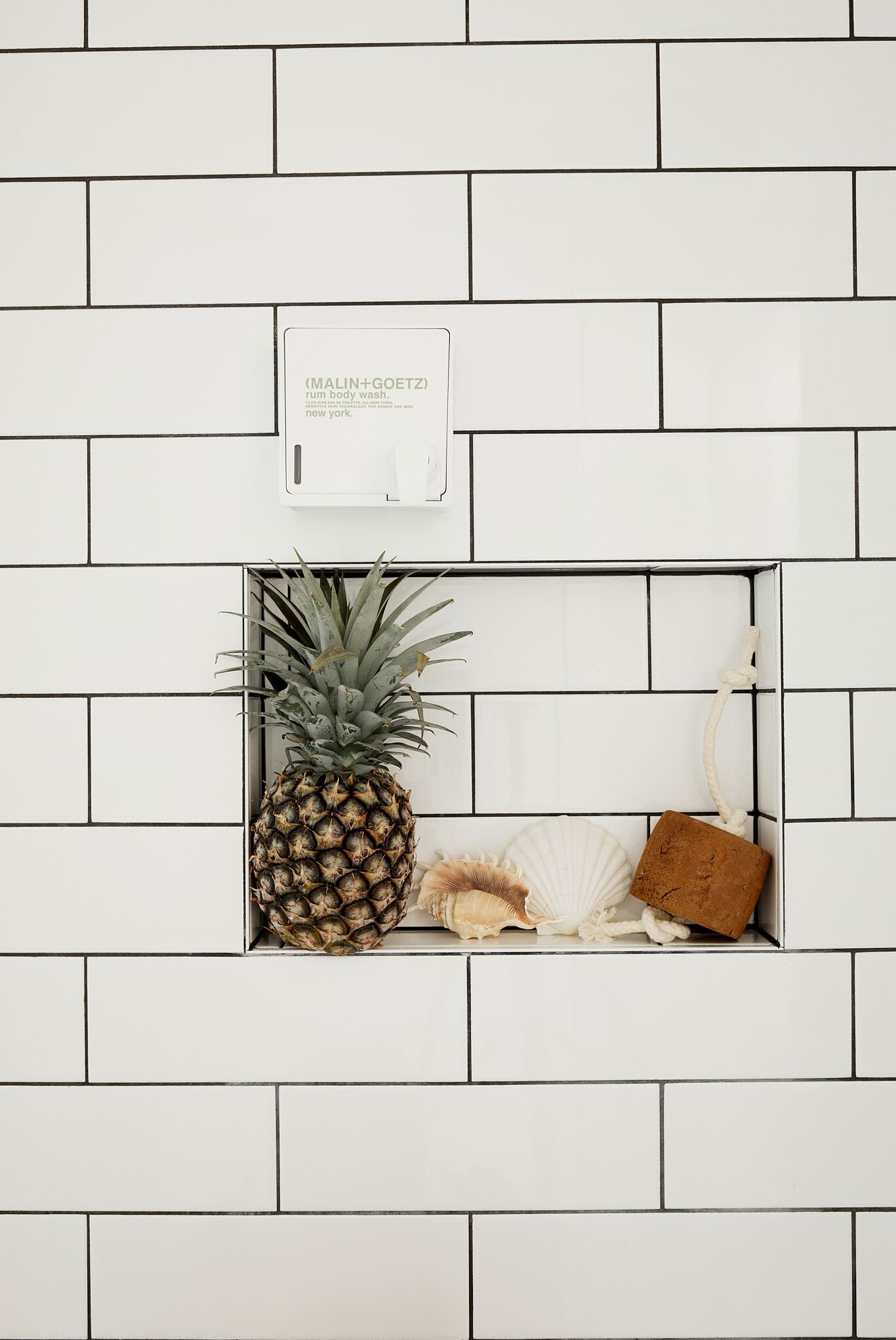 A white tiled wall with a small shelf containing a pineapple, a seashell, and a brown object.