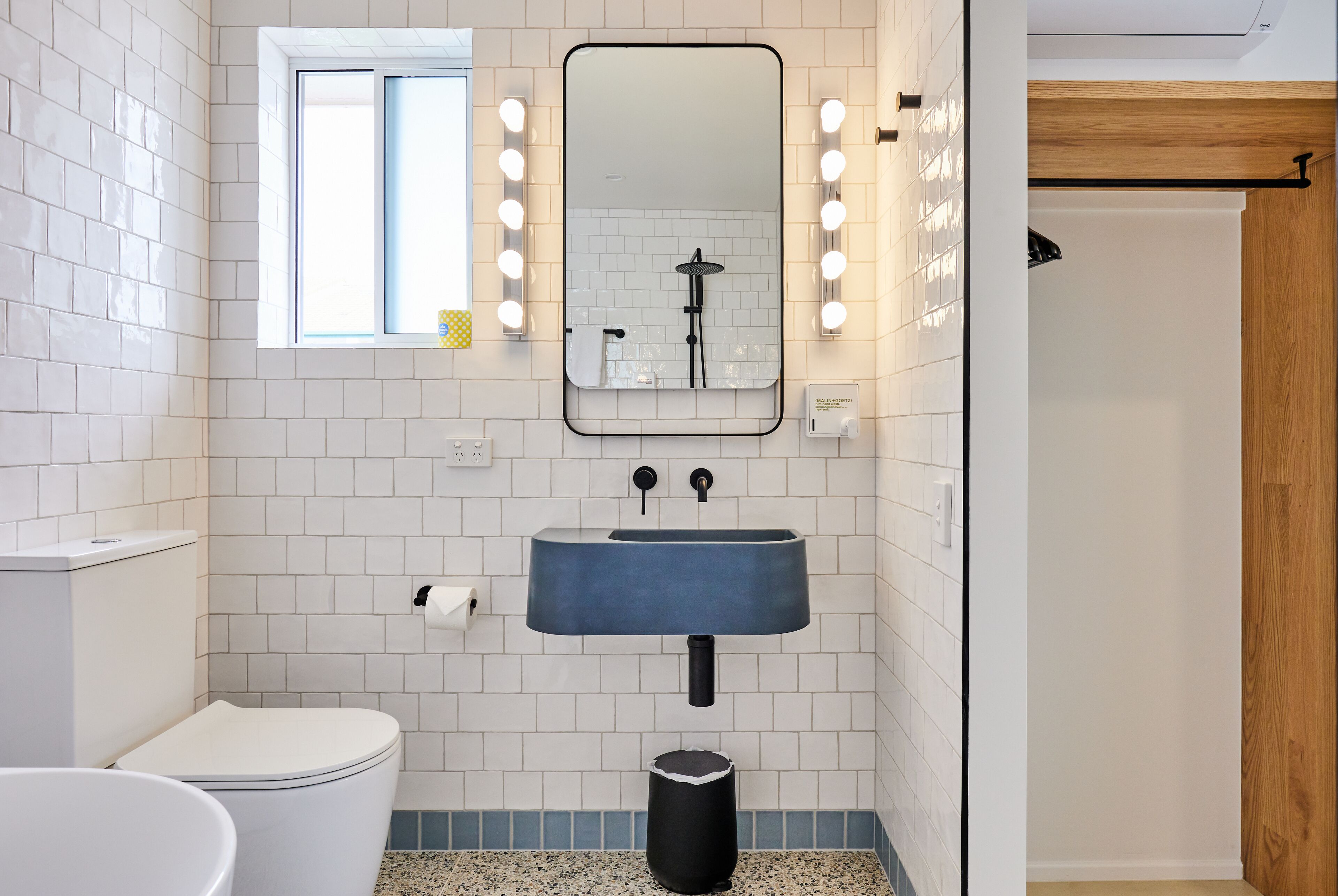 Een badkamer met een witte betegelde muur, een blauwe wastafel, een spiegel met verlichting, een toilet en een prullenbak.