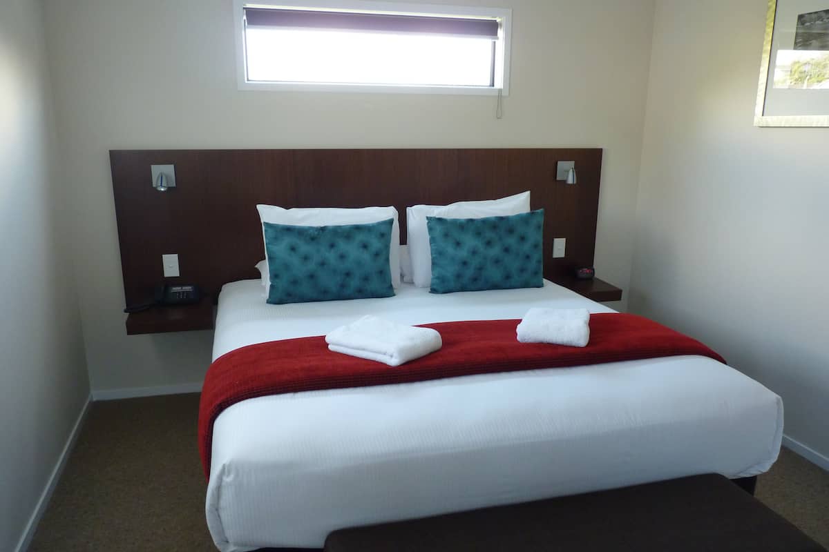 Cama bem arrumada com lençóis brancos, duas almofadas verde-água e um cobertor vermelho. Há uma mesinha de cabeceira com um abajur e um telefone.