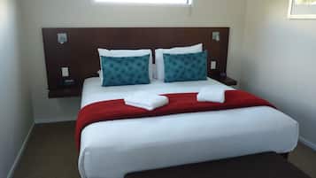 Una cama bien hecha con sábanas blancas, dos almohadas color turquesa y una manta roja. Hay una mesita de noche con una lámpara y un teléfono.