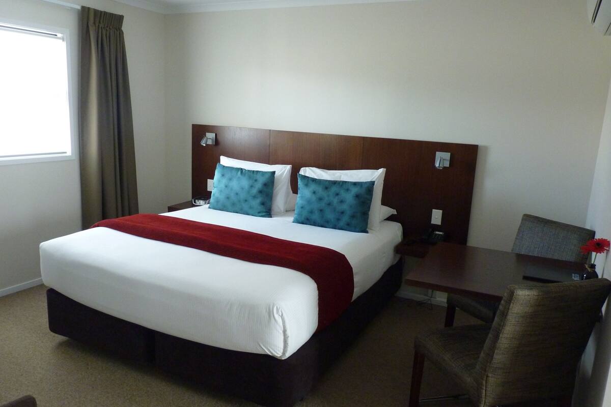 Quarto de hotel com uma cama grande, duas almofadas, um cobertor vermelho, cabeceira de madeira, mesa de cabeceira com telefone, uma cadeira e uma janela com cortinas.