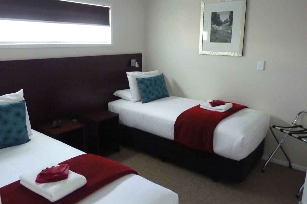 Quarto de hotel com duas camas de solteiro, cada uma com colchão branco, manta vermelha e travesseiro azul. Uma pequena mesa de cabeceira com um controle remoto. Um quadro emoldurado na parede acima das camas.