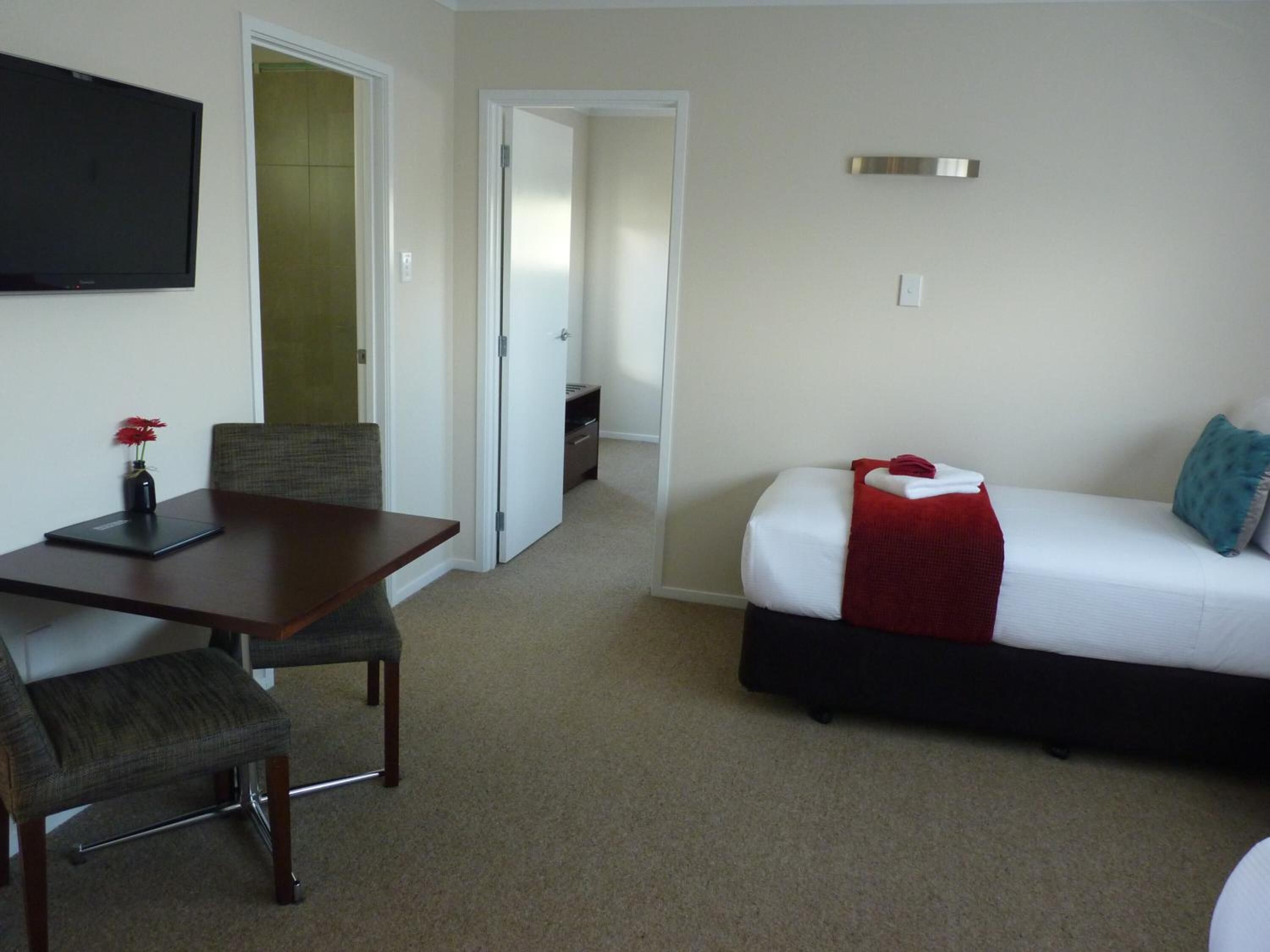 Habitación de hotel con cama, escritorio y silla. Hay un televisor montado en la pared y una puerta que da a otra habitación.