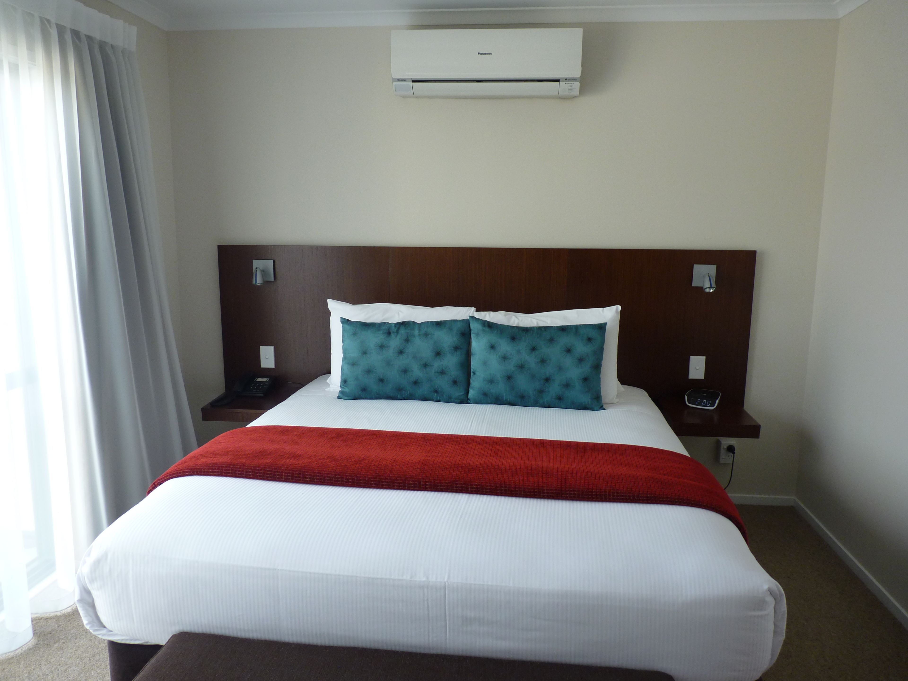 A hotel room with a large bed, a red throw blanket, two blue pillows, a white headboard, and an air conditioning unit mounted on the wall.