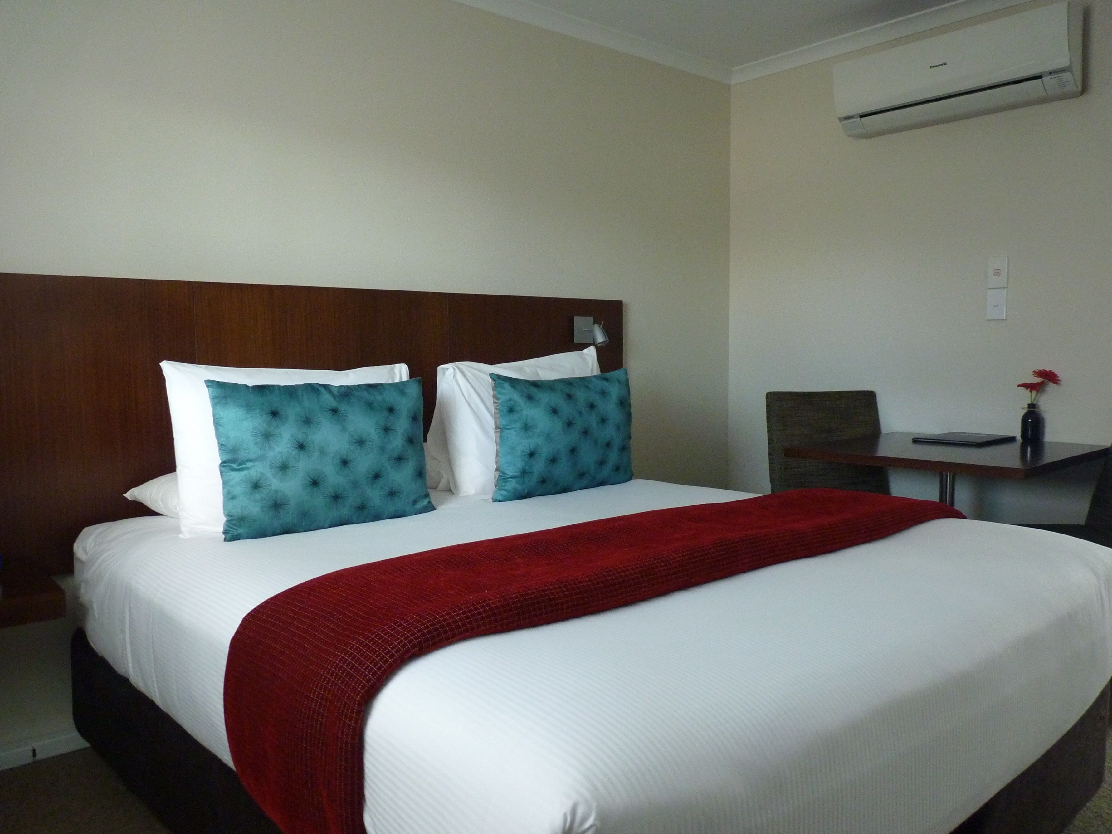 Habitación de hotel con una cama grande, dos almohadas, una manta roja, cabecera de madera, una mesita con un jarrón de flores y un equipo de aire acondicionado en la pared.