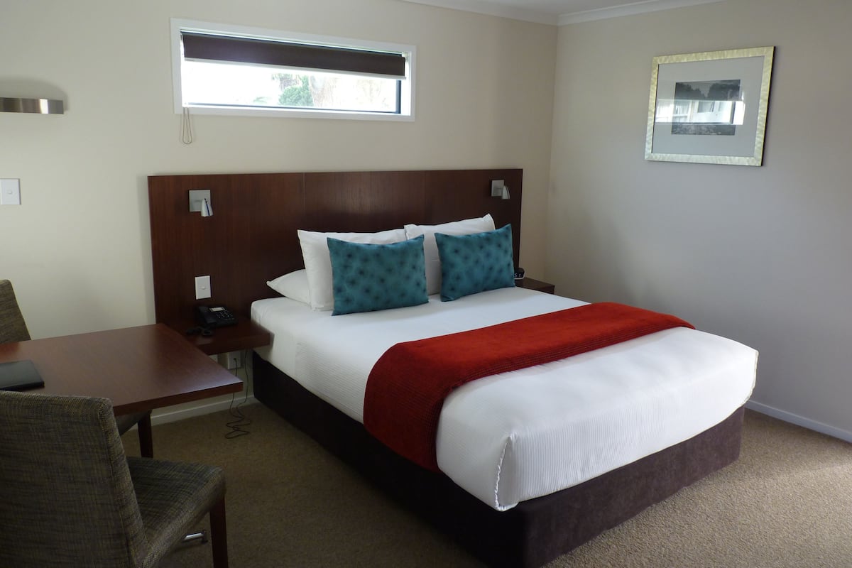 Quarto de hotel com uma cama grande, cabeceira de madeira, escrivaninha com cadeira, mesa de cabeceira, janela e um quadro na parede.