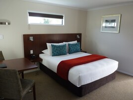 Habitación de hotel con una cama grande, cabecera de madera, un escritorio con silla, mesita de noche, ventana y un cuadro enmarcado en la pared.