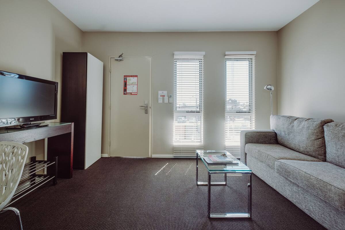 A modern living room with a flat-screen TV, a glass coffee table, a sofa, and a chair. There is a door with a bulletin board and a window with blinds.
