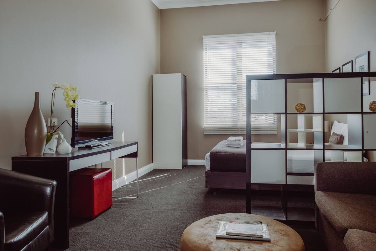 A modern living room with a flat-screen TV, a sofa, a red ottoman, and a large window with blinds.