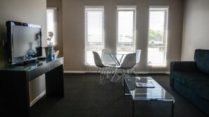 A modern living room with a flat-screen TV, a glass coffee table, and a sofa. There are three windows with blinds, and a chair is placed near the windows. - Crossroads Hotel Narrabri (Narrabri)