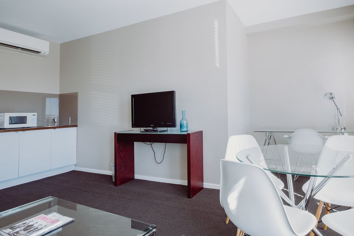 A modern hotel room with a flat-screen TV on a wooden desk, a glass dining table with white chairs, a microwave, and a small blue vase.