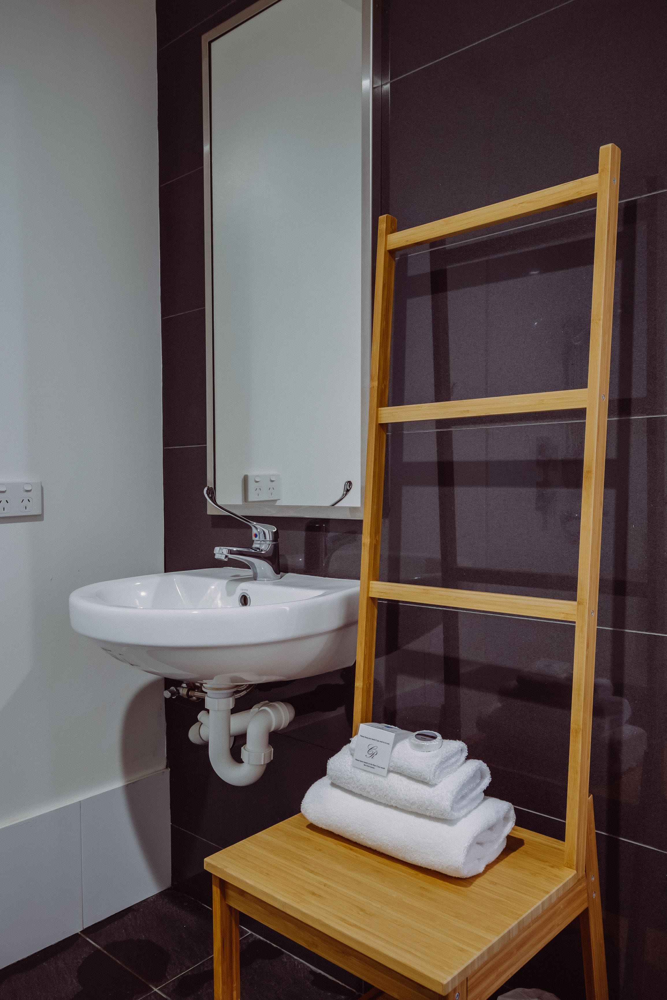 Une salle de bain avec un lavabo blanc, un miroir et une étagère en bois où sont pliées des serviettes.