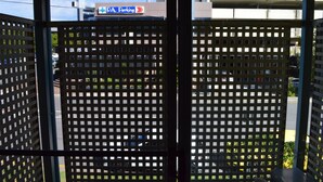 Visão pela janela, com grade de metal, placa indicando área de estacionamento e um edifício ao fundo.