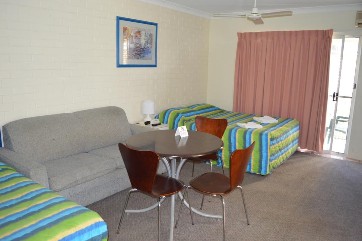 A room with a grey sofa, a round table with four chairs, a bed with striped bedding, and a ceiling fan.