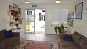 Uma área de recepção com balcão, placa de 'Não há quartos disponíveis', placa de recepção, planta e janela com persianas.