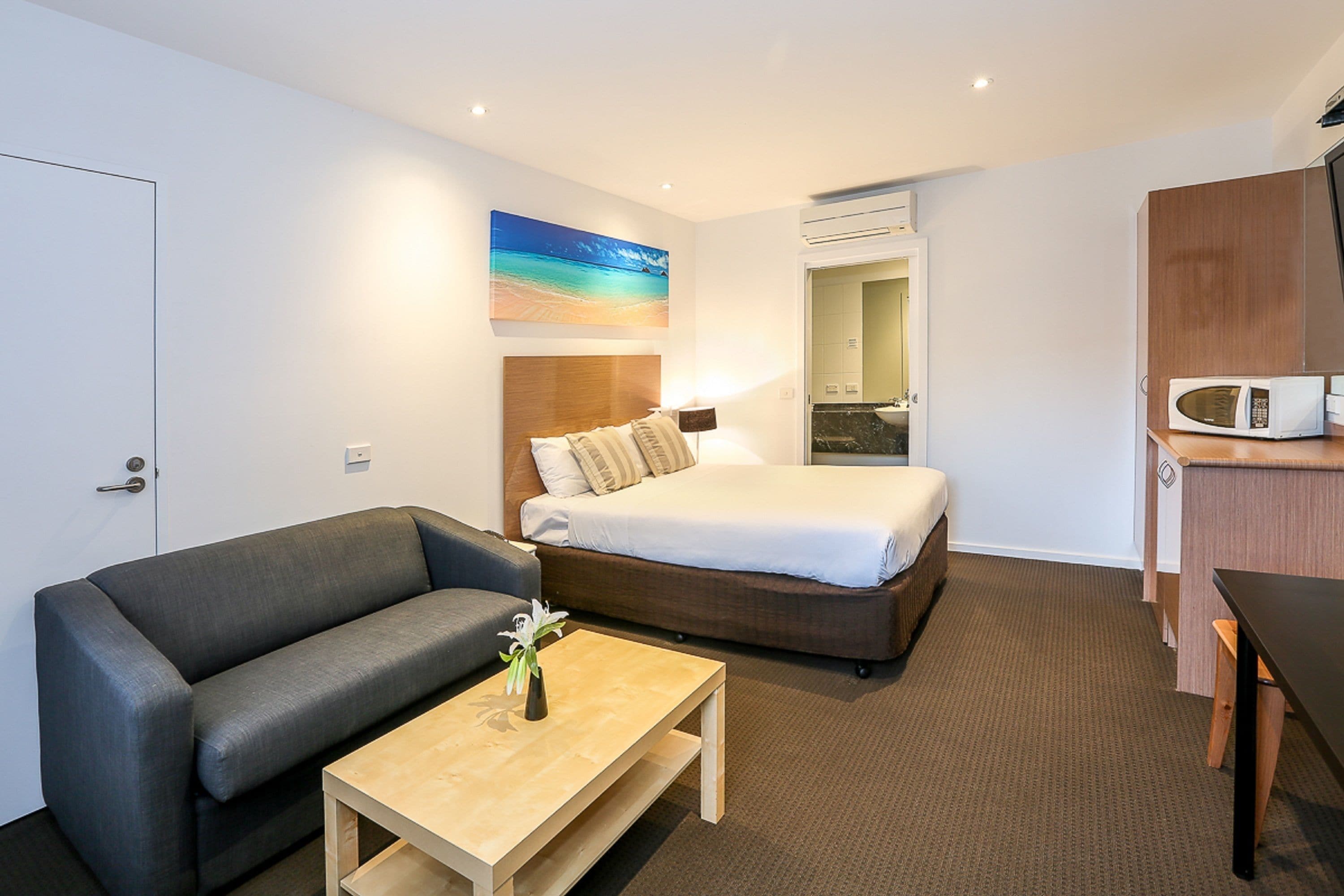Quarto de hotel com uma cama, um sofá, uma mesinha com um vaso, micro-ondas e um banheiro visível por uma porta aberta.