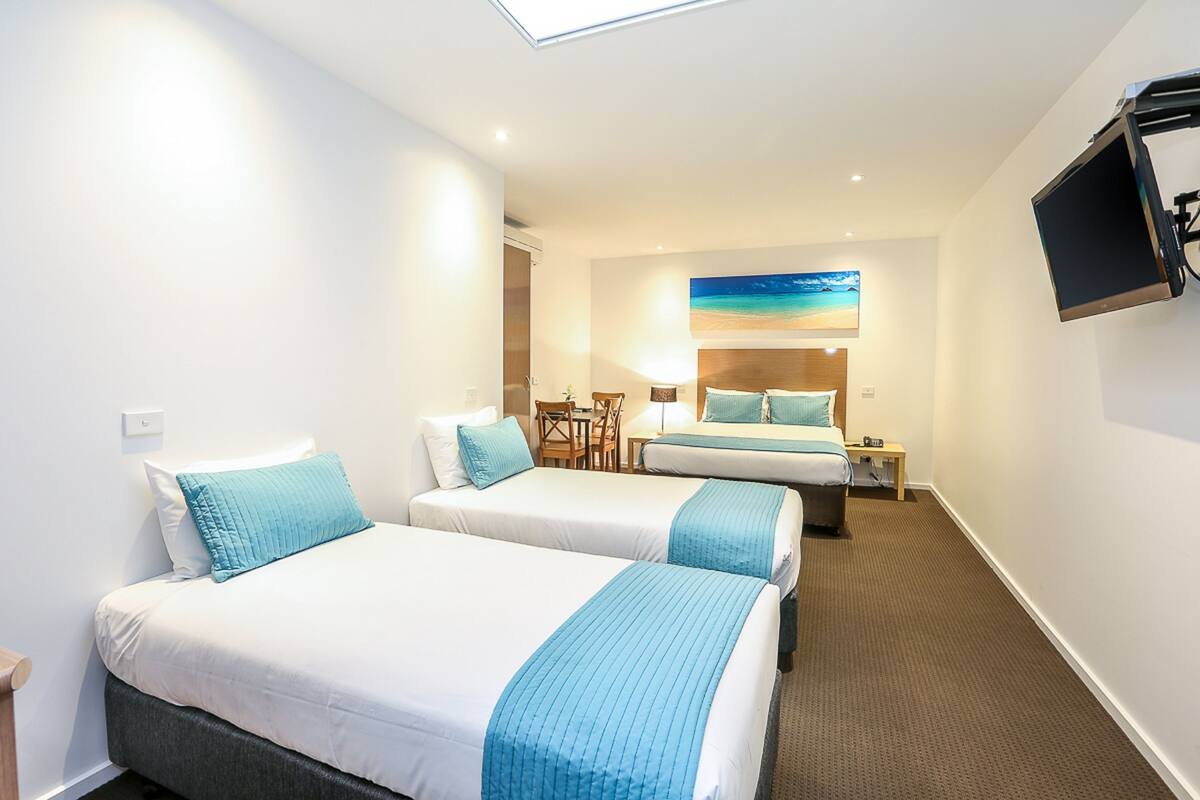 A hotel room with two beds, a TV mounted on the wall, a desk with a chair, and a painting of a beach scene.
