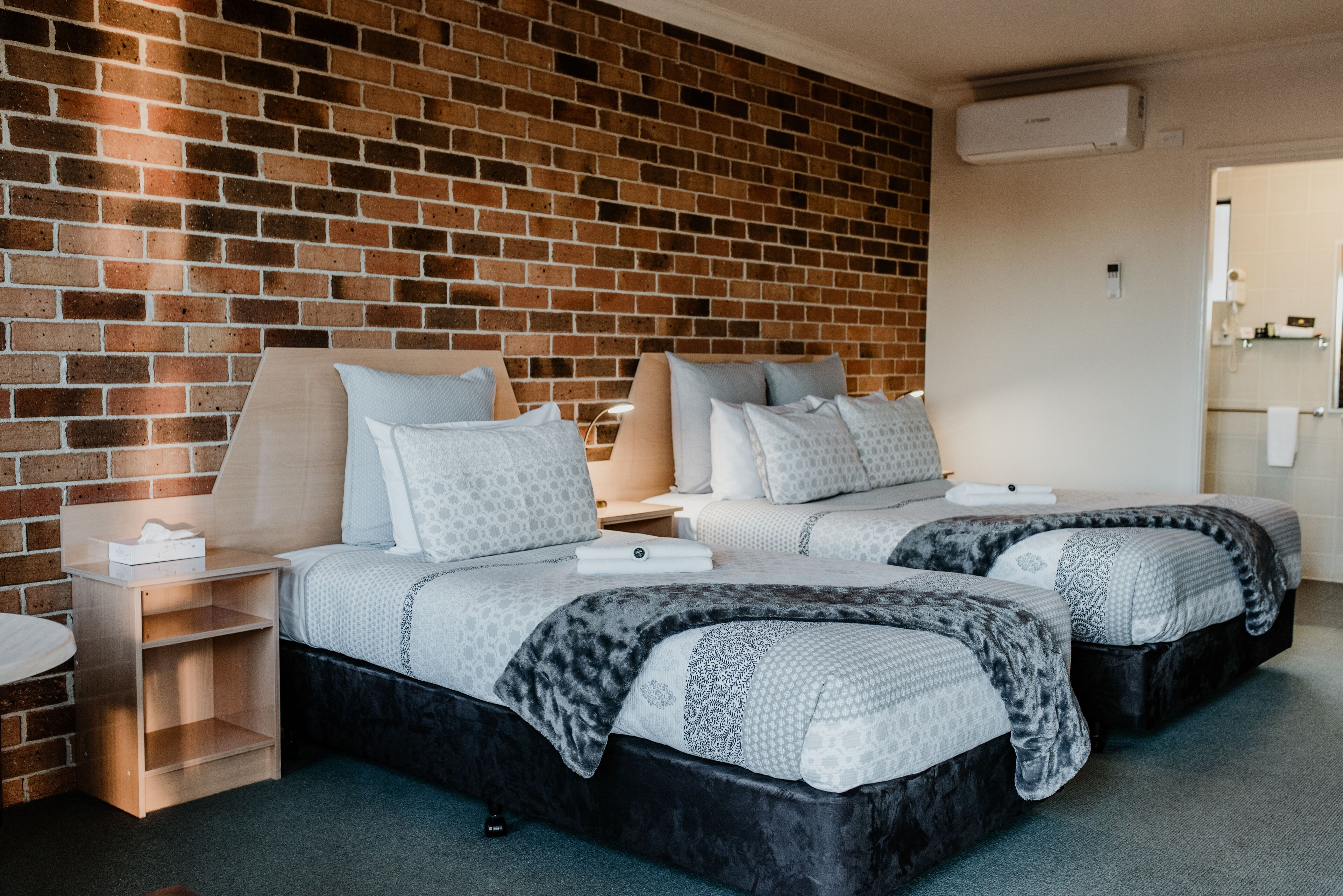 Ein Hotelzimmer mit zwei Betten, einer Ziegelwand, einem kleinen Nachttisch und einem durchgegangene Tür erkennbaren Badezimmer.
