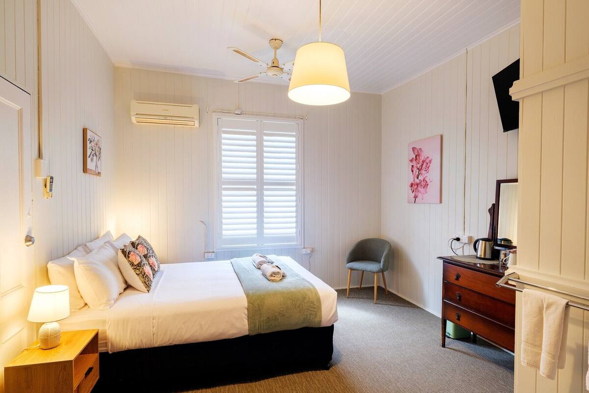 A bedroom with a bed, bedside table, lamp, chair, dresser, and a window with blinds.