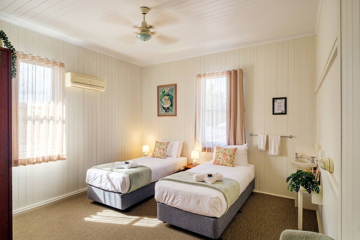 A hotel room with two beds, a ceiling fan, a window with curtains, a bedside table with a lamp, and a small plant.