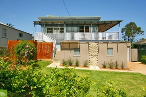 Une maison à deux étages dotée d’un balcon, d’une façade en pierre et d’un petit jardin devant, avec une clôture en bois et des plantes en fleurs.