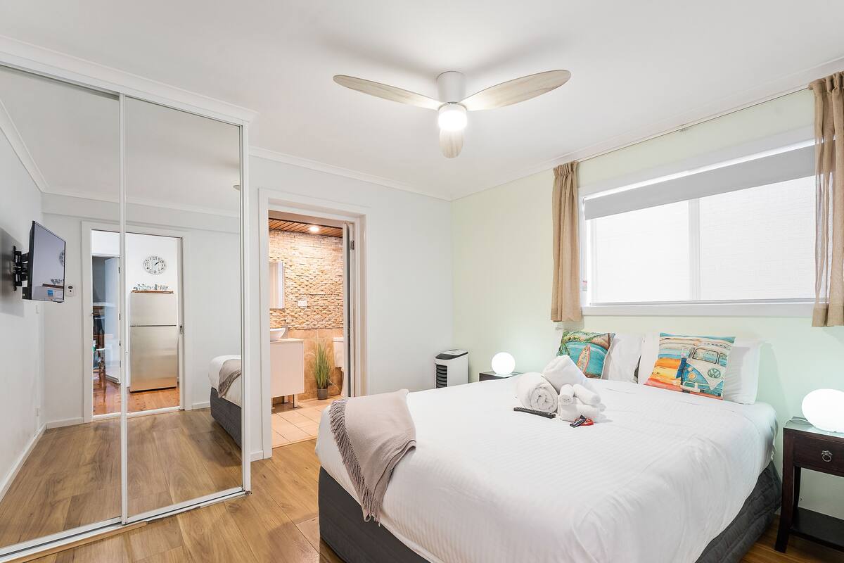 Un dormitorio con cama, mesitas de noche, ventilador de techo, ventana con cortinas, espejo y baño visible a través de una puerta abierta.
