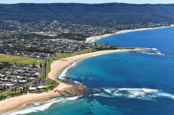 An aerial view of a coastal town with a beach, residential area, and forested hills.