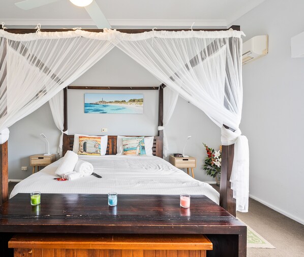 Un dormitorio con cama, mesitas de noche, lámpara, reloj y planta en la mesita, además de un cuadro en la pared.