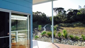 A modern house with a glass door leading to a deck, surrounded by a landscaped garden with a pathway and small plants. - a Dune Escape (Emu Bay)