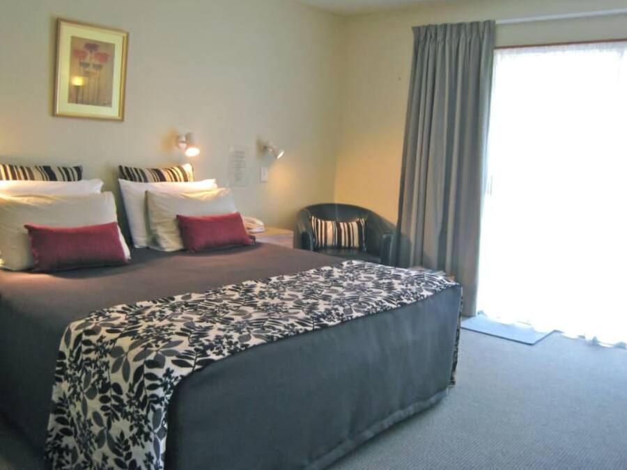 A hotel room with a large bed, two bedside tables, a chair, a window with curtains, and a wall-mounted lamp.