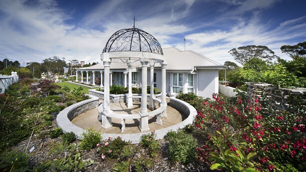 A white gazebo with a dome roof surrounded by a circular stone base, set in a landscaped garden with various plants and flowers. - Katoomba Manor (Katoomba)