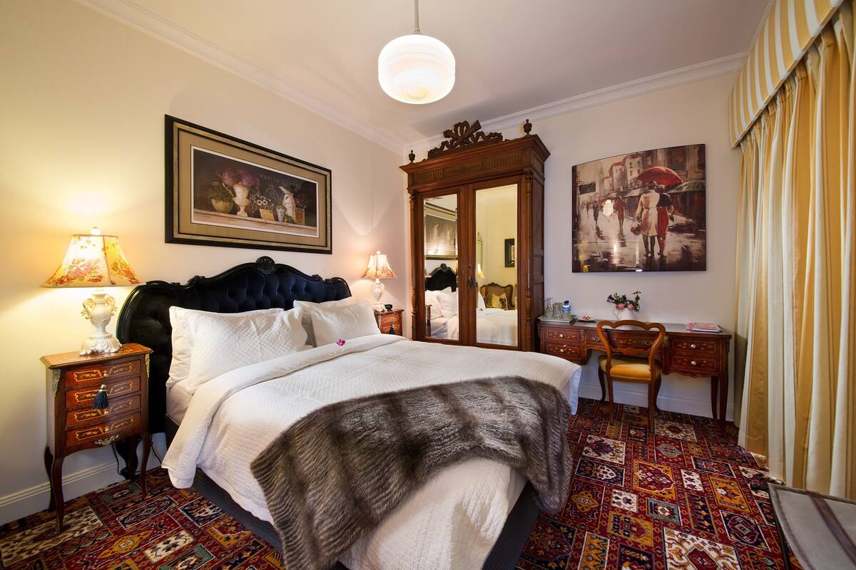 A bedroom with a large bed, two bedside tables with lamps, a chair, a desk, a mirror, and two framed artworks on the walls.
