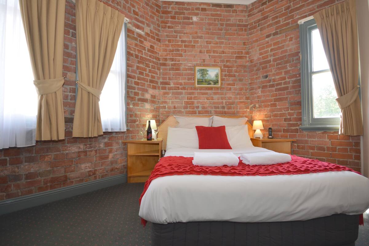 A brick-walled bedroom with a large bed, two bedside tables with lamps, and a window with curtains.