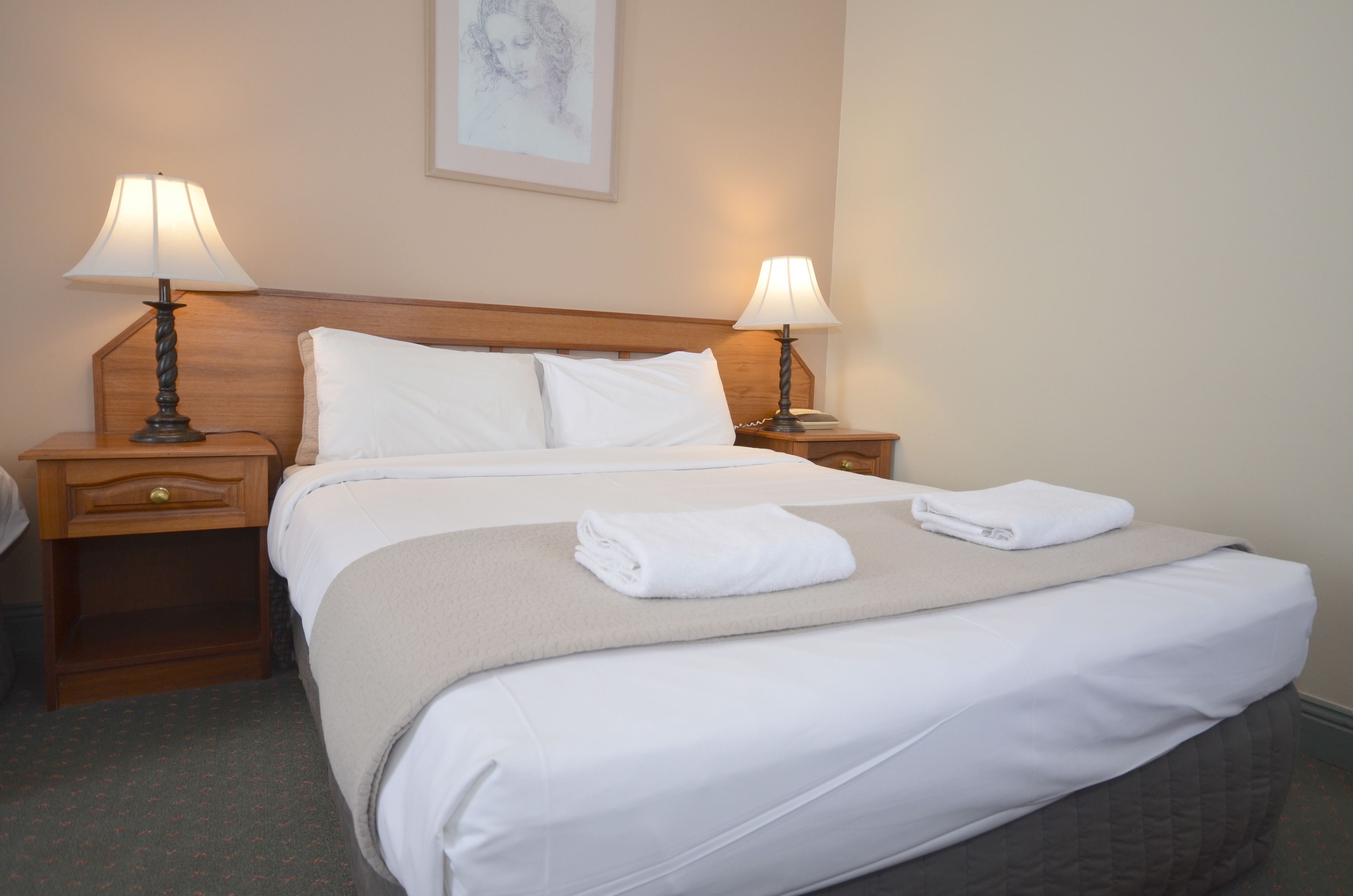 A neatly made bed with white linens and a beige throw blanket, flanked by two bedside tables with lamps, in a hotel room.