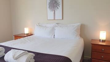 A neatly made bed with white linens, two bedside tables with lamps, a framed picture of a palm tree on the wall, and a purple quilted bedspread.