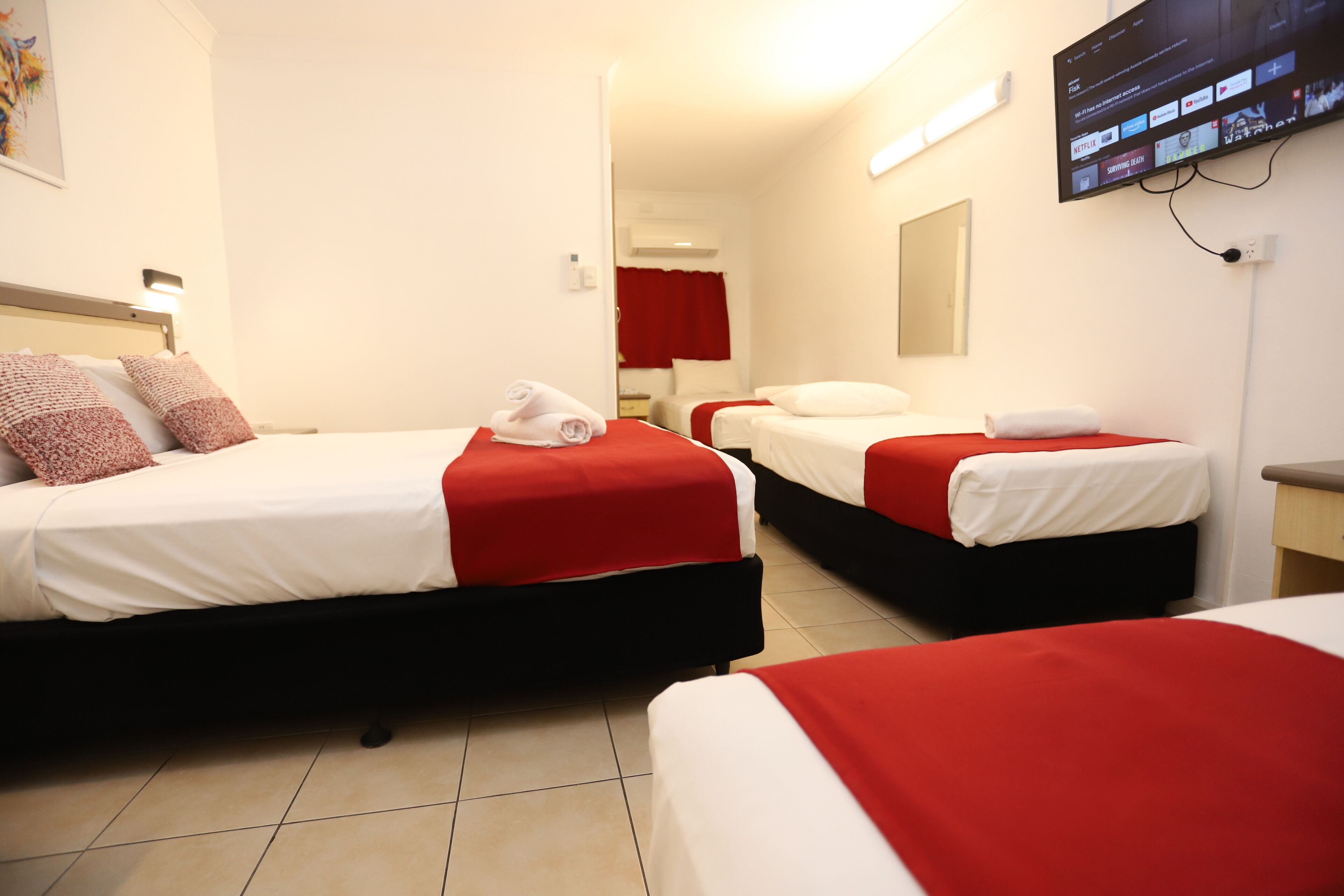 A hotel room with three beds, each with red and white bedding, a flat-screen TV mounted on the wall, and a red curtain in the background.