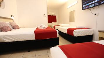 A hotel room with three beds, each with red and white bedding, a flat-screen TV mounted on the wall, and a red curtain in the background.